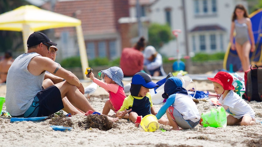 A man and woman sit on the sand at Williamstown beach with four kids wearing hats playing in the sand.