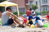 A man and woman sit on the sand at Williamstown beach with four kids wearing hats playing in the sand.