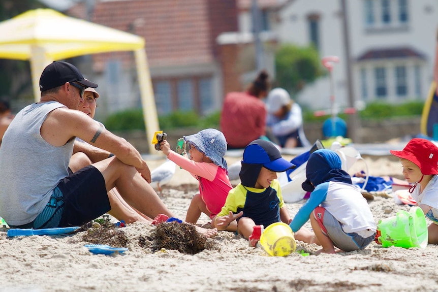 A man and woman sit on the sand at Williamstown beach with four kids wearing hats playing in the sand.