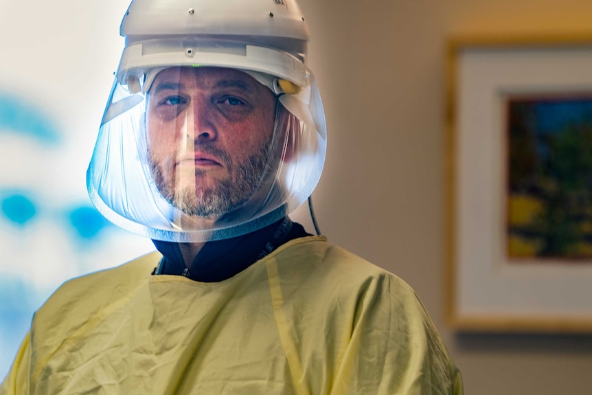 A bearded man looking solemn in a face shield and yellow hospital gown