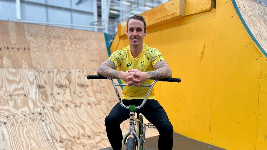 A man in a green and gold jerset sits on a BMX bike smiling at the camera