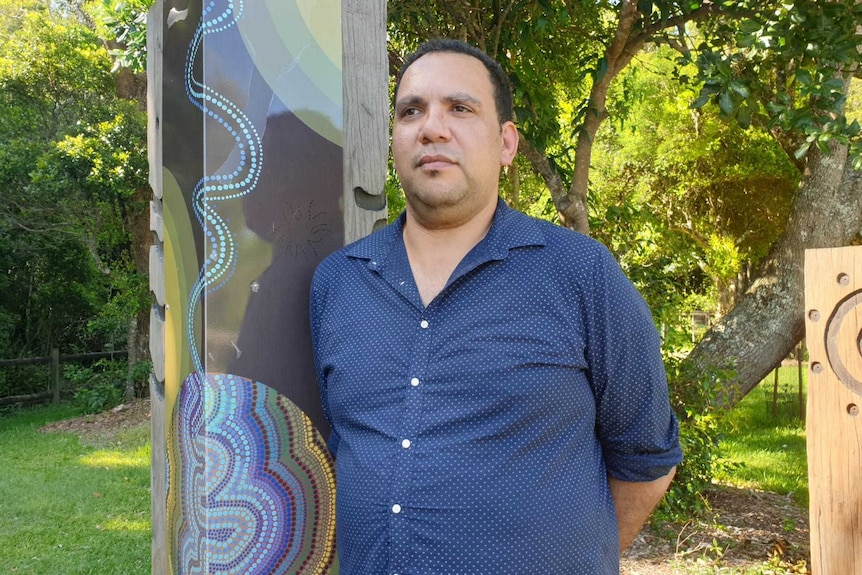 An Indigenous man stands in a leafy area with his hands behind his back.