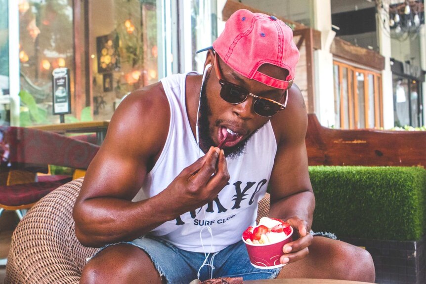 A man in a tanktop and backways cap eating cake and holding ice cream