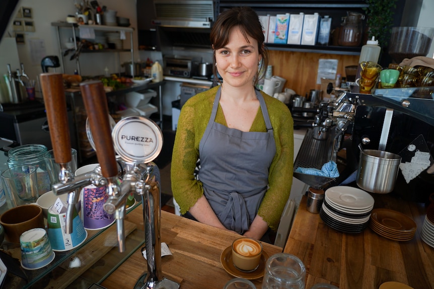 A woman in a green top with a denim apron stands behind the counter of a cafe