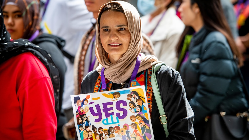 A smiling woman in a hijab holds up a sign that says 'Yes' for the Voice to Parliament.