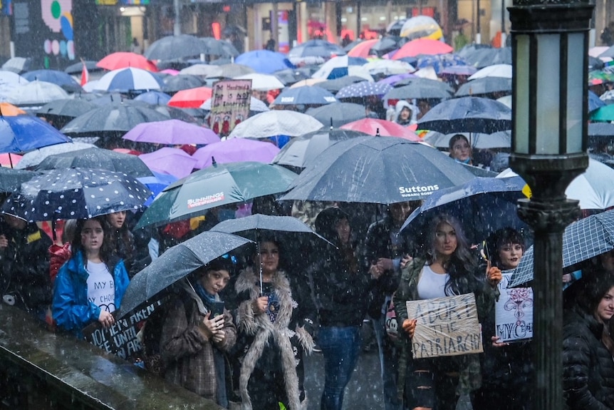 A crowd stands under a gloomy sky and rain with umbrellas.