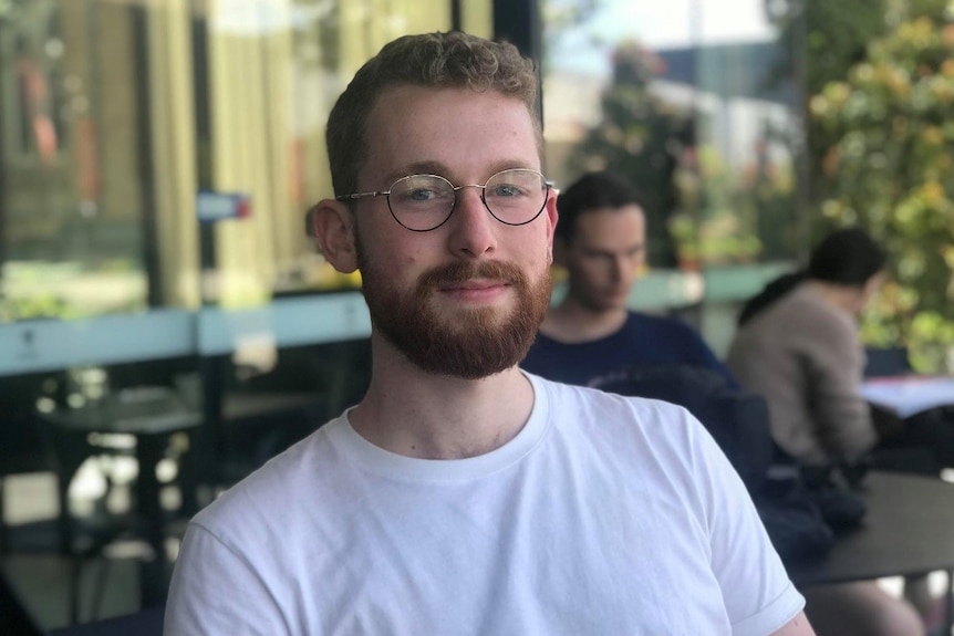 A young man wearing glasses looks into the camera against a leafy, cafe background.