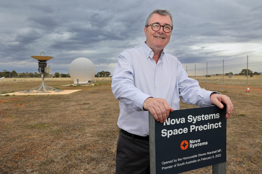 A man with glasses and greying hair places his hands on a sign reading "Nova Systems Space Precinct".