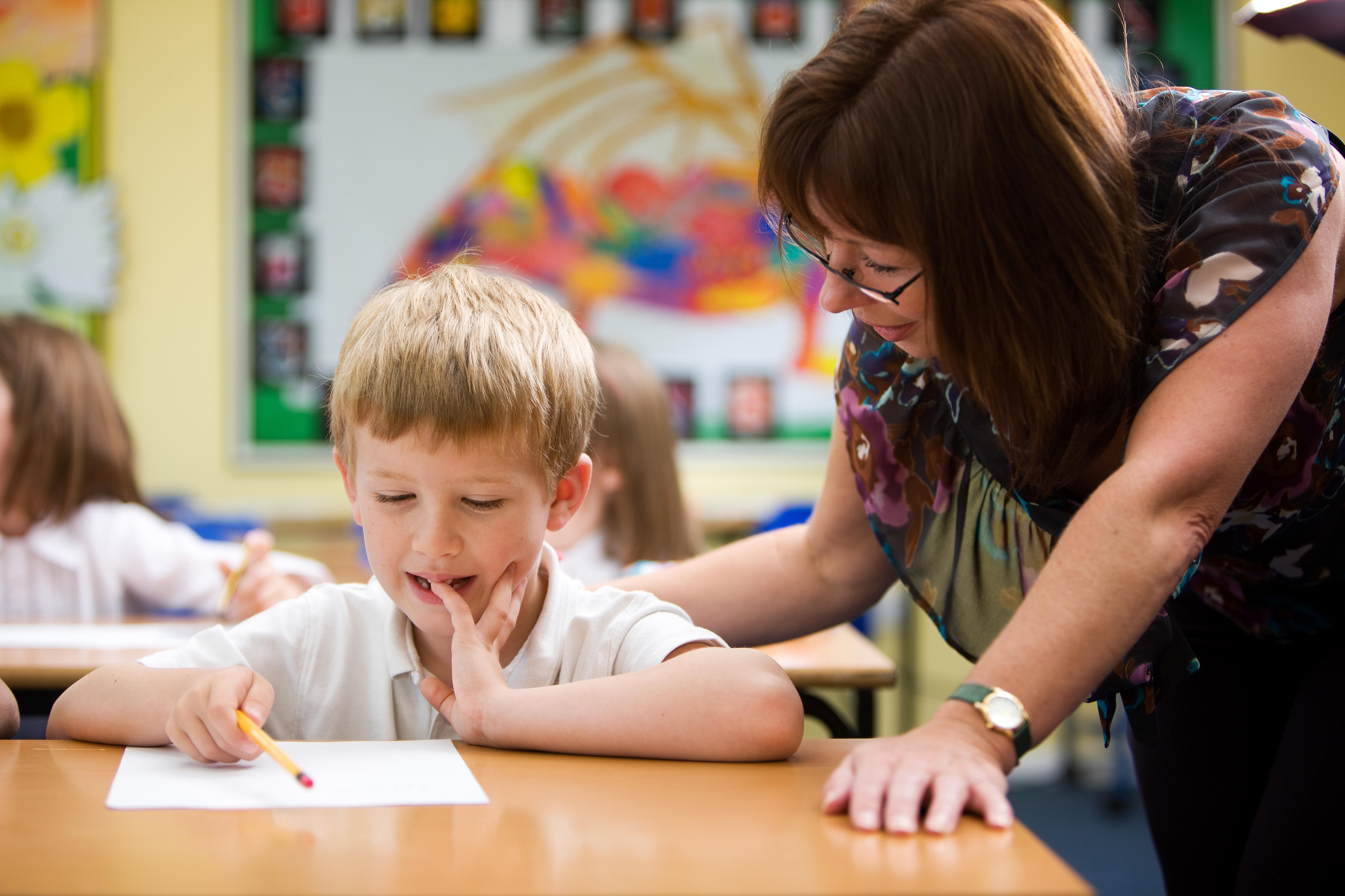 A teacher leans over her primary student’s desk to help him with a maths problem. He is thinking, with his finger in his mouth.