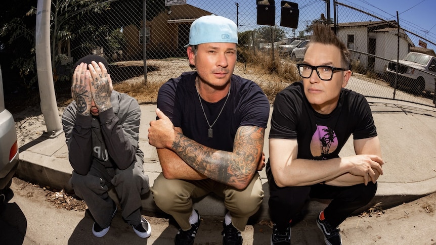 The three members of Blink-182 sit on a kerb in a neighbourhood in the sun