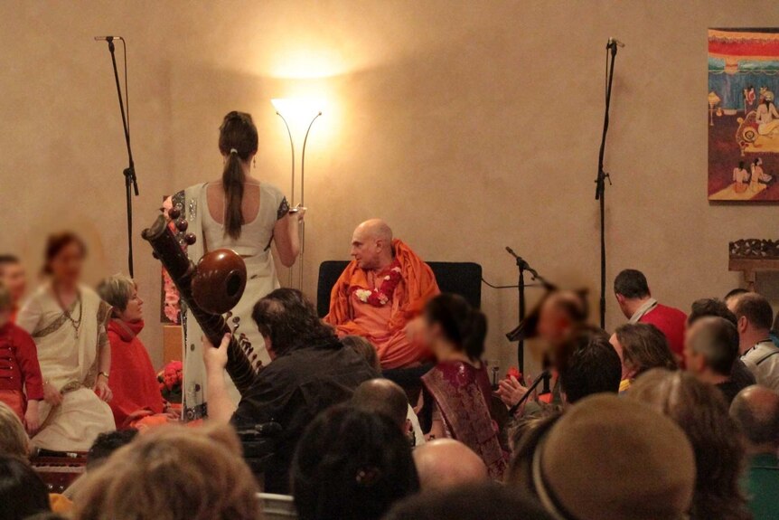 Russell Kruckman is shown dressed in orange sitting on a chair surrounded by followers seated on the floor.