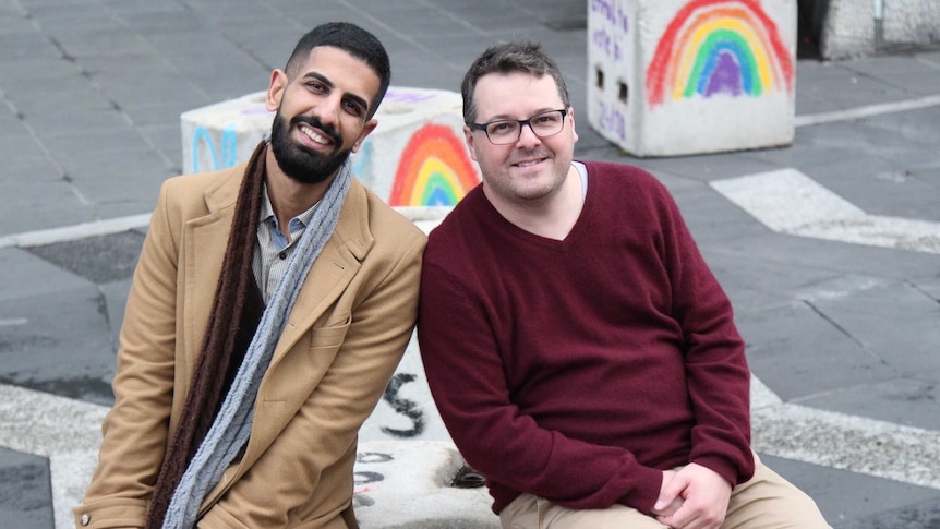 Two men sit on a concrete bollard and smile at the camera. Two bollards in the background have rainbows painted on them.