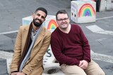 Two men sit on a concrete bollard and smile at the camera. Two bollards in the background have rainbows painted on them.
