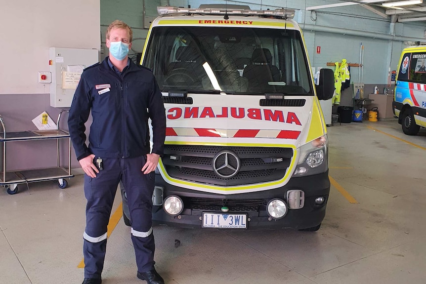 A tall man with blond hair stands in front of an ambulance in a big shed