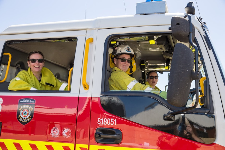 Three women wearing yellow hi-vis sit in a red fire truck smiling out the window 