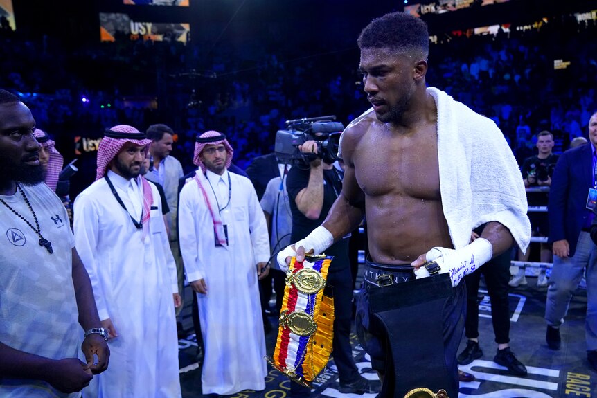 Anthony Joshua holds two boxing belts while draped with a white towel as a line of men look on