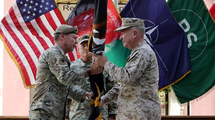 A man in a US military uniform hands a rolled up flag to another man.