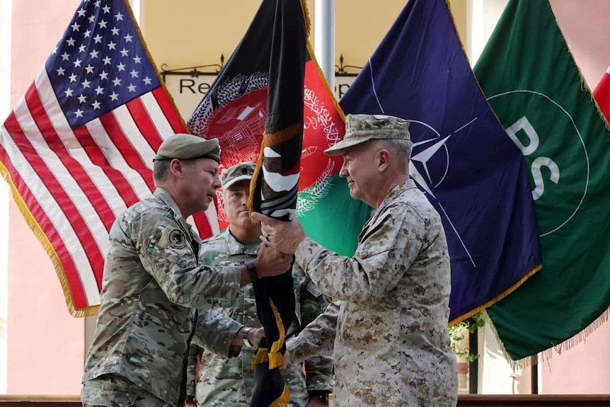 A man in a US military uniform hands a rolled up flag to another man.
