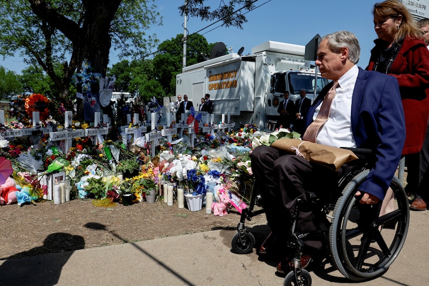 A white man with grey hair in a suit wheels himself past masses of flowers, candles and white crosses bearing names