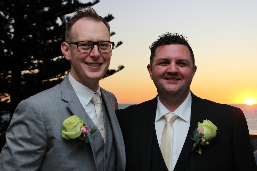 Two men smile, they have short hair wear a grey and black suit with white roses.