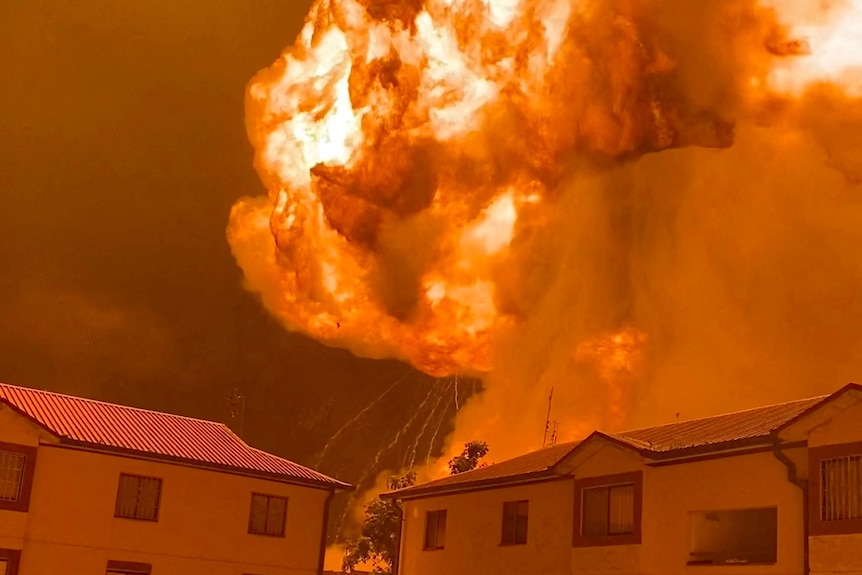 A huge ball of flame erupts into the night sky behind two small blocks of units and a small carpark.
