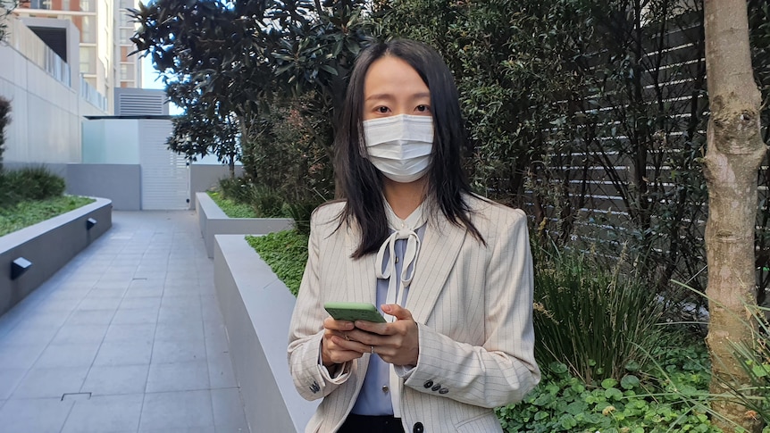 A woman wearing a mask stands outside an apartment development.