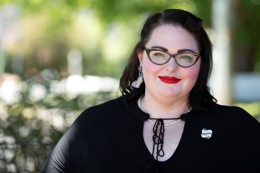 A woman with black hair, black glasses and red lipstick looks at the camera and smiles. 