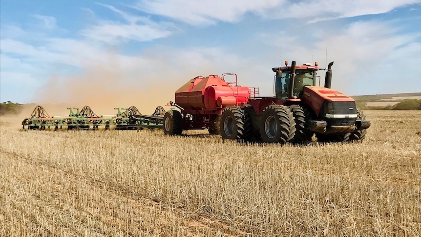 A red tractor pulls a seeding rig through stubble. The sky is blue with some faint cloud. Dust swirls behind the seeder 