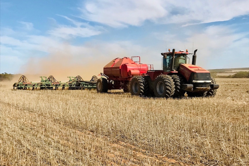 A red tractor pulls a seeding rig through stubble. The sky is blue with some faint cloud. Dust swirls behind the seeder 