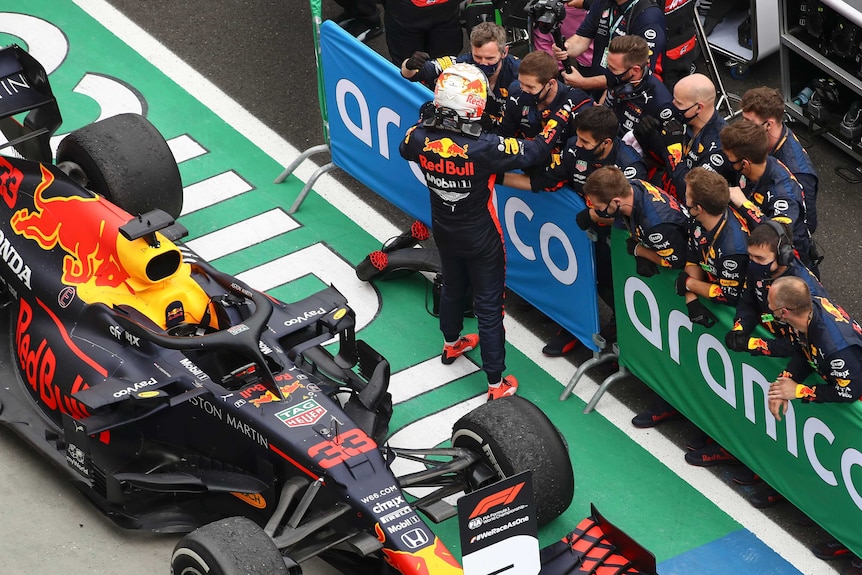 Max Verstappen stands in front of a group of men wearing his full race kit including helmet with his car behind him