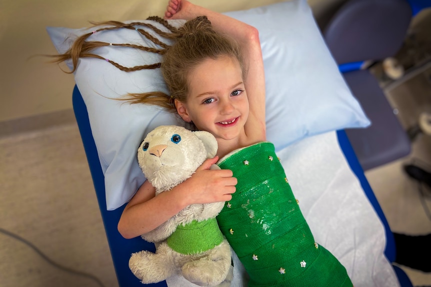 A young girl lays on hospital bed with green body cast with stars, she holds a white teddy bear with a matching green cast.