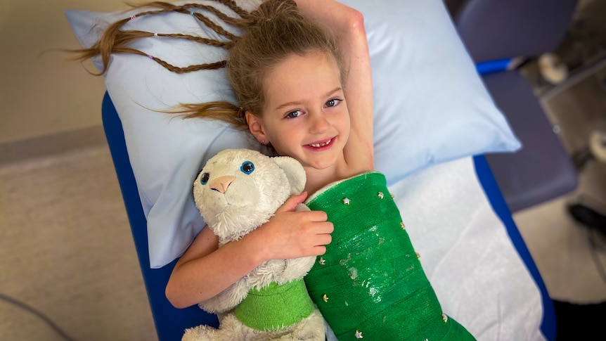 A young girl lays on hospital bed with green body cast with stars, she holds a white teddy bear with a matching green cast.