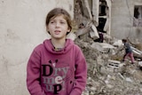 A close up of a young girl wearing a pink jumper standing in front of rubble.