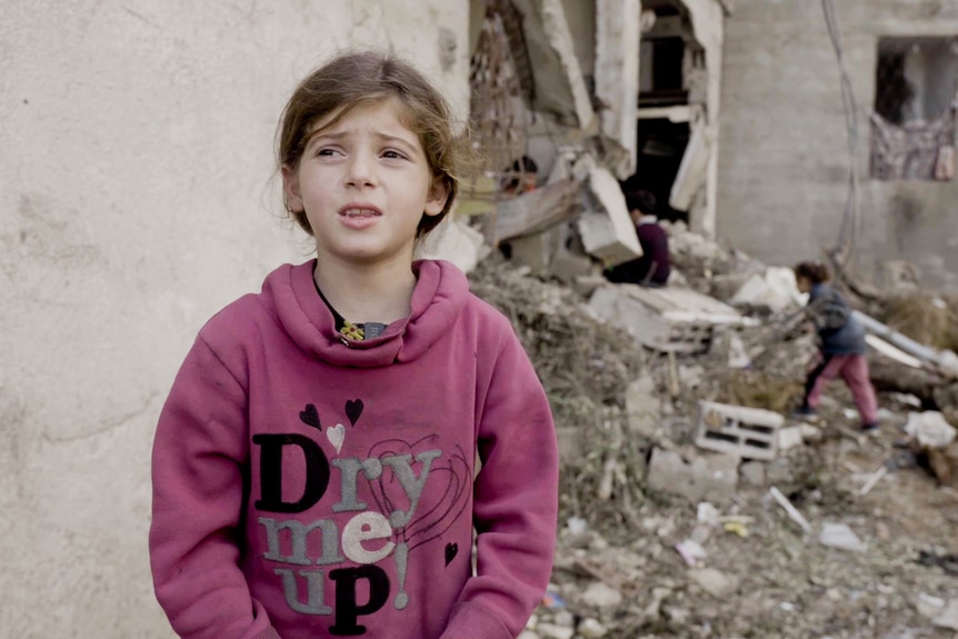 A close up of a young girl wearing a pink jumper standing in front of rubble.