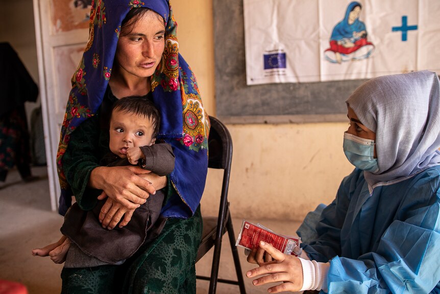 A woman sitting holding a baby next to another woman sitting holding a small package both in head scarfs