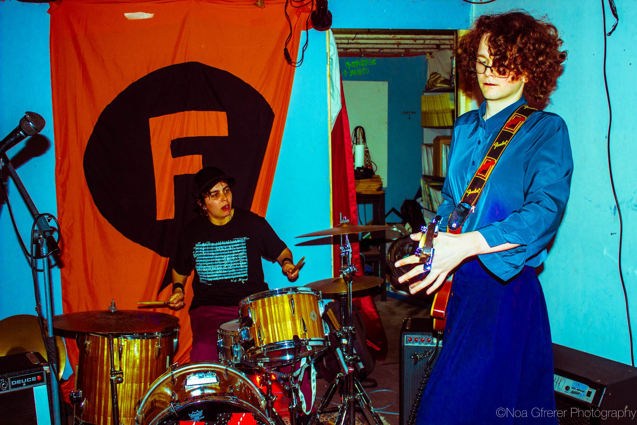 Young woman plays guitar with drummer in background.