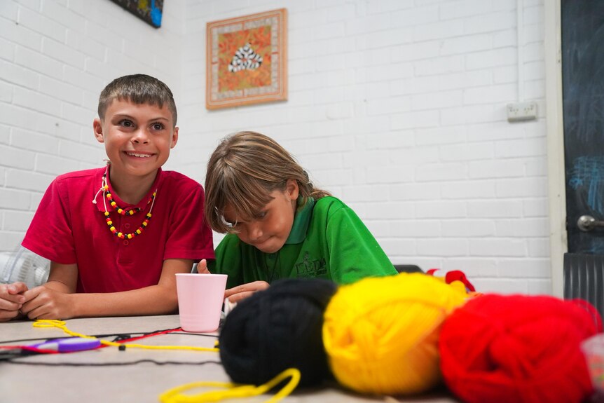 A boy wearing a red shirt smiling at the camera and a girl wearing a green shirt playing with thread. Red, yellow and black thre