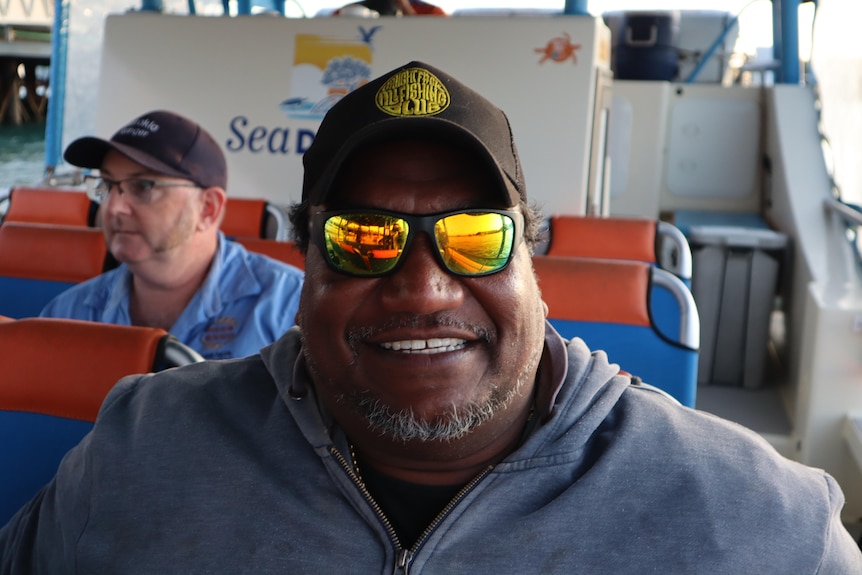 Man wearing hat and sunglass smiles into the camera while sitting on a seat on a ferry