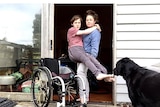 A woman lifts a young man out of his wheelchair