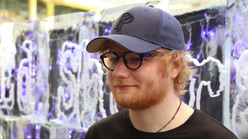 Musician Ed Sheeran wearing a cap and glasses talks to media with a large ice sculpture in the background.