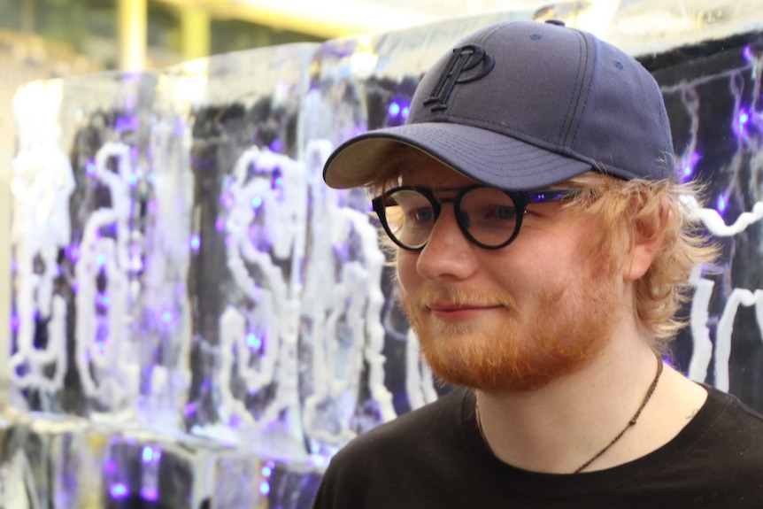 Musician Ed Sheeran wearing a cap and glasses talks to media with a large ice sculpture in the background.