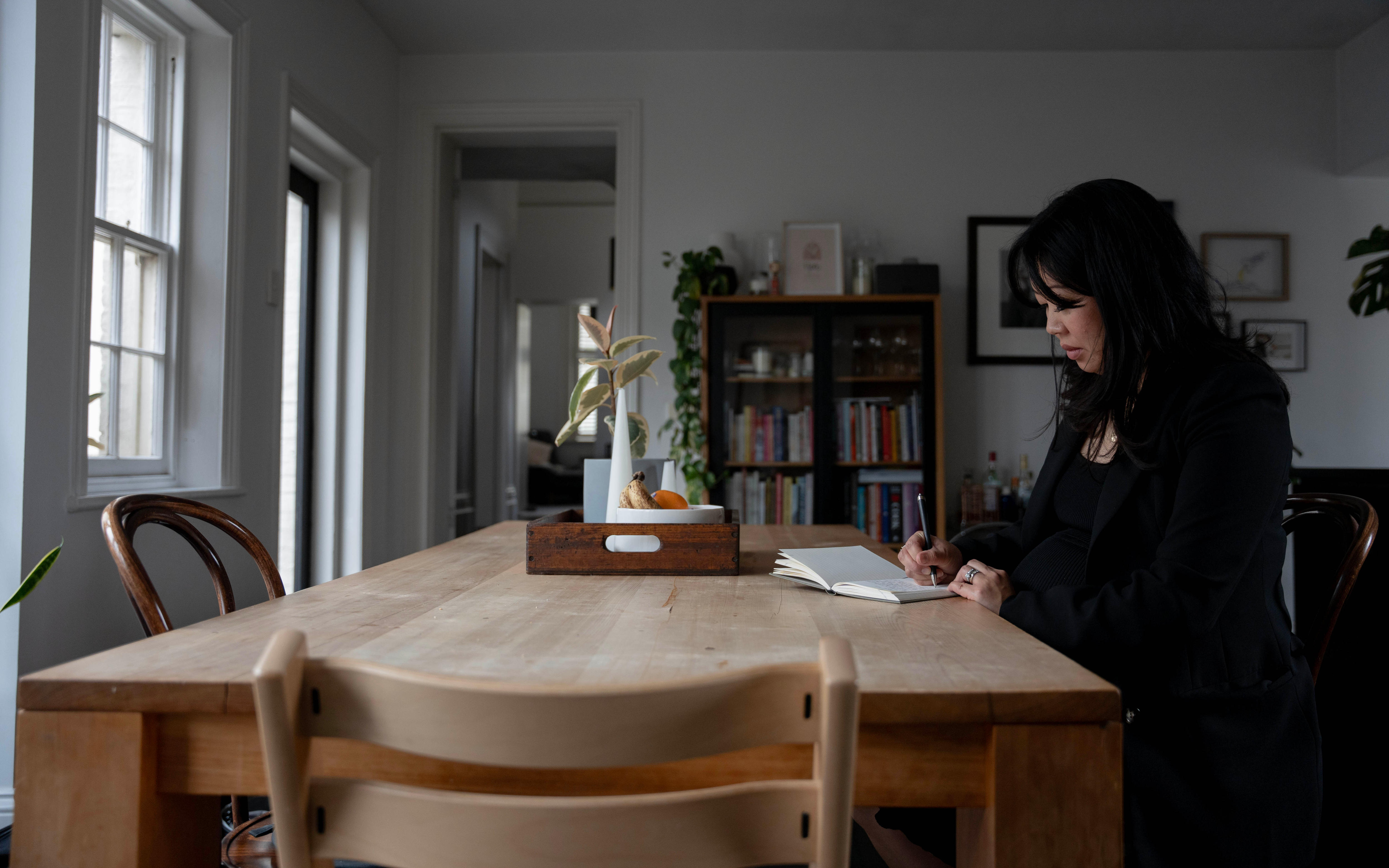 A woman with black hair sits at a table and writes in a book