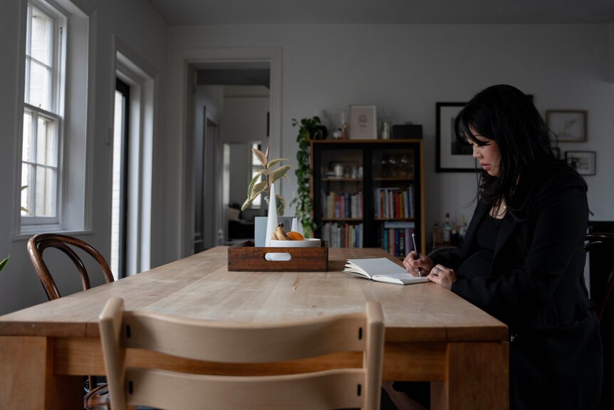 A woman with black hair sits at a table and writes in a book