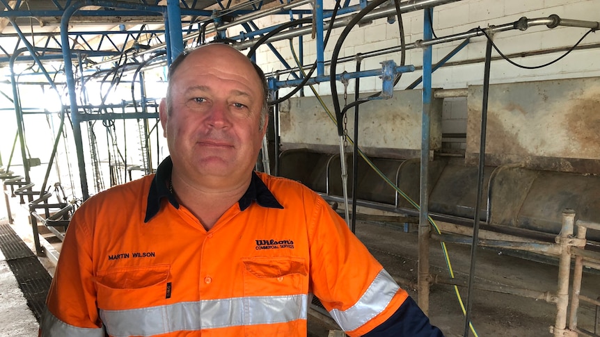A man in a high vis orange long sleeved shirt stands in a dairy shed with no cows in it.