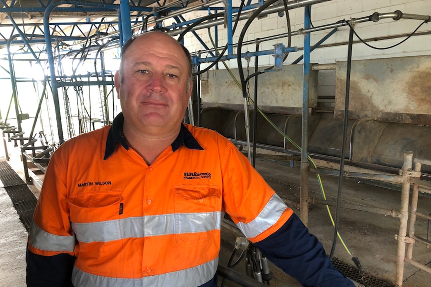 A man in a high vis orange long sleeved shirt stands in a dairy shed with no cows in it.