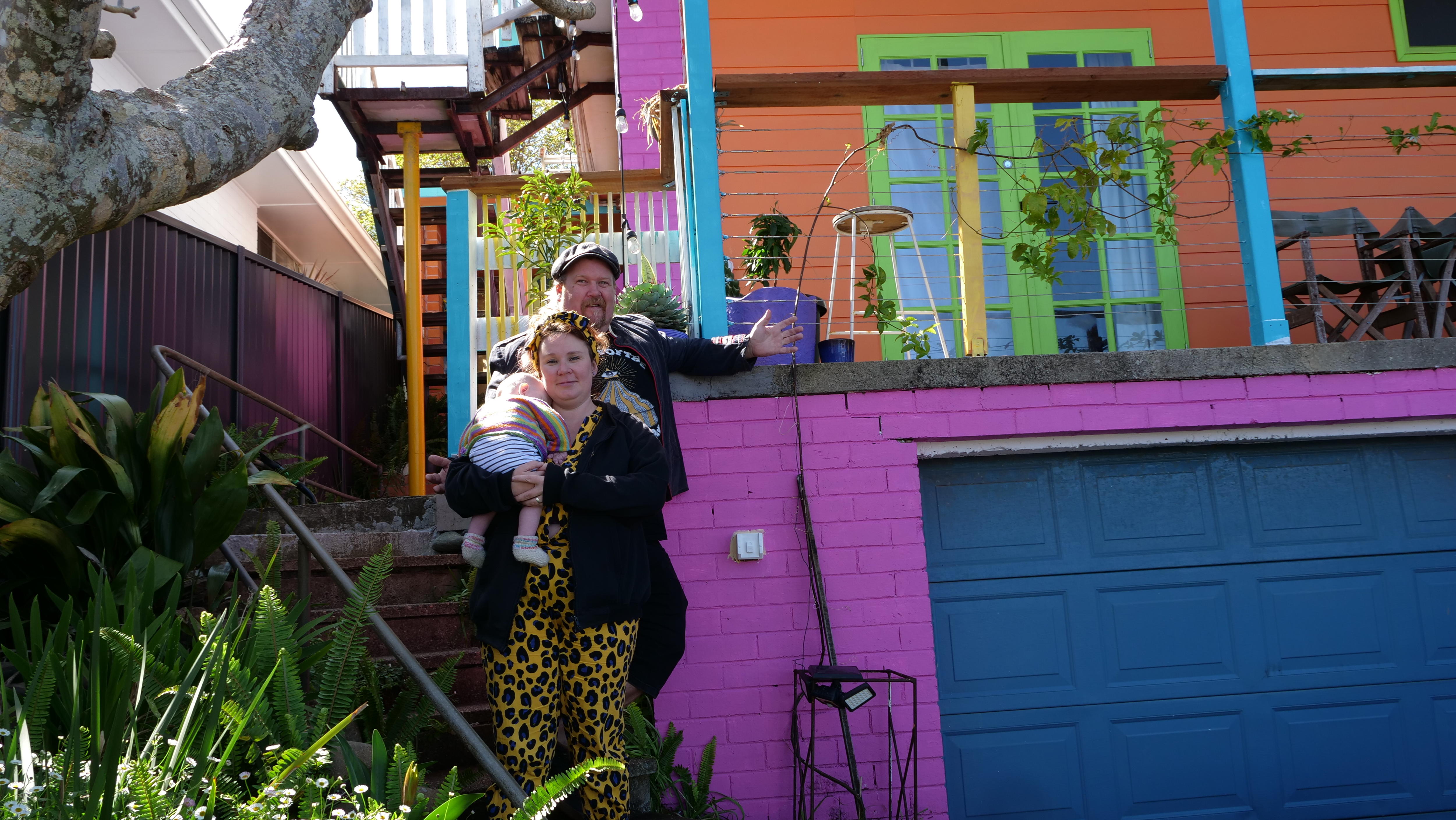 A couple and a baby, mum in yellow leopard print, on the steps of their house painted pink, orange, blue and green.