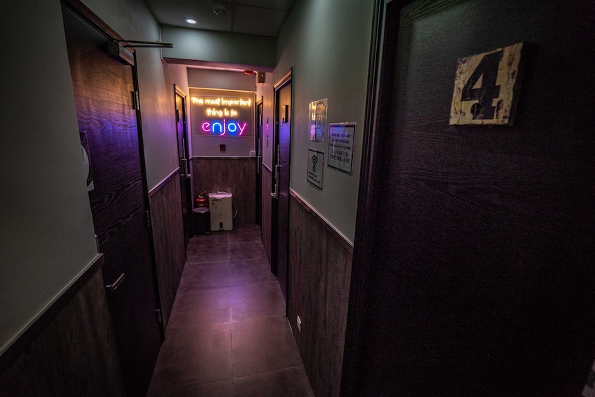 A dark hotel hallway with black, numbered doors on either side and a neon sign that says "the most important thing is to enjoy".