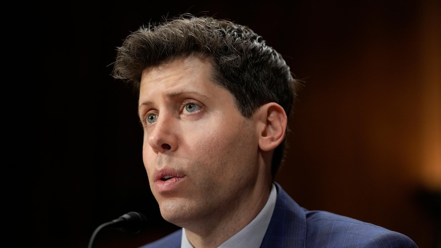 A white man with short hair speaks into a small desk microphone, while wearing a suit and tie