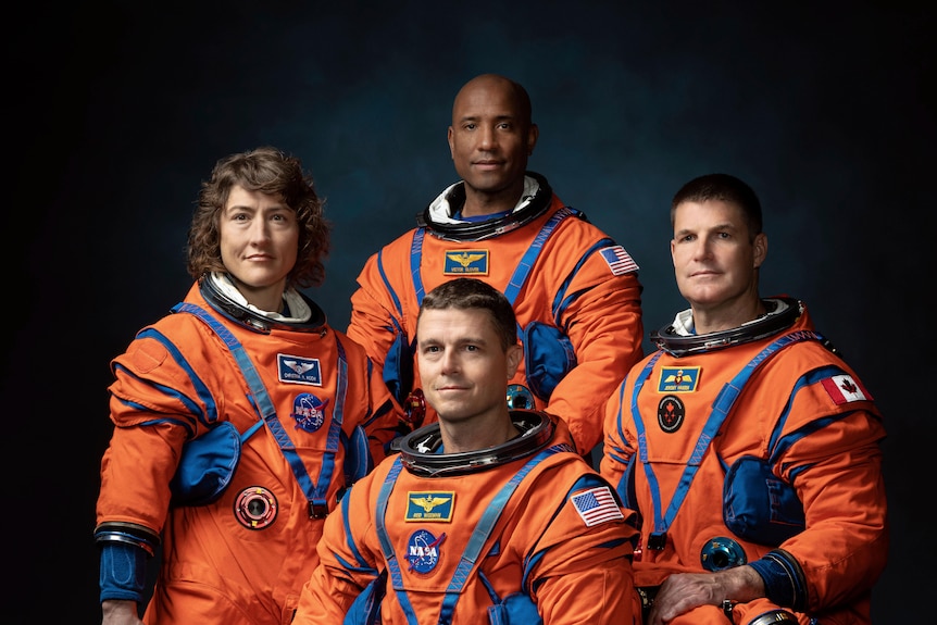 The four astronauts pose for a photo wearing orange suits. 