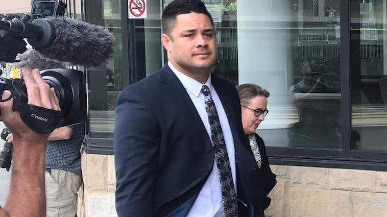 A man aged in his 30s, wearing a dark suit, walks down a walkway in front of a court building while being filmed.